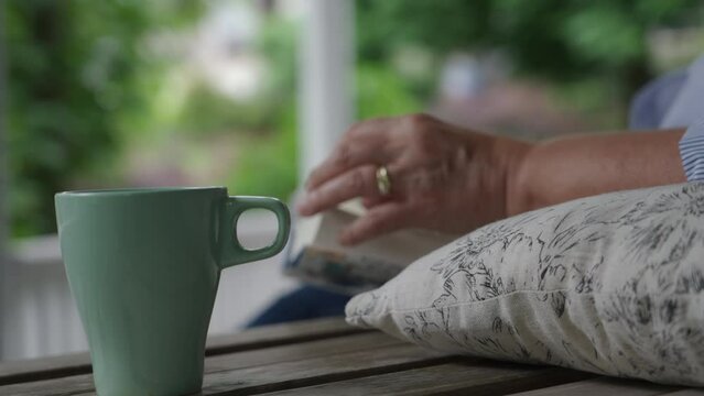 Close up of green coffee or tea mug with the hands of a woman, in soft focus, wearing wedding rings and reading a book, on a comfortable, cozy outdoor veranda with a white railing and trees   