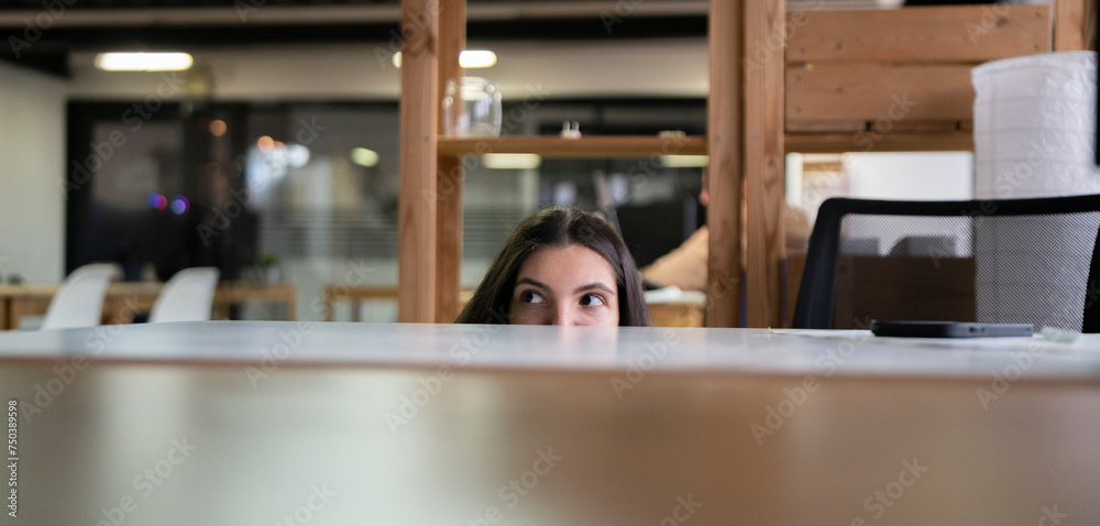 Wall mural office worker sneaking a peek above laptop in a modern workspace during daytime, hiding from boss