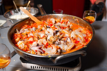 Seafood Paella Cooking in a Large Pan. A large pan filled with seafood paella featuring shrimp and squid, simmering on a stove, with the vibrant colors of fresh ingredients on display.
