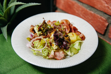 Potato Salad with Crispy Bacon Strips on Plate. A hearty plate of potato salad garnished with crispy bacon strips, red onion, and fresh lettuce, served on a textured white plate.