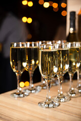 Champagne Glasses on Table at Festive Event. Rows of sparkling champagne glasses on a wooden table, with the warm bokeh of lights in the background at a festive celebration.