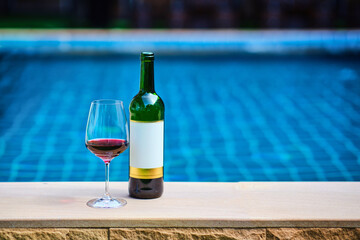 A bottle of red wine and a glass of wine are set on the edge of a clear blue pool, surrounded by a...