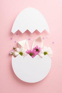 Dive into our lavish Easter arrangement. Overhead vertical picture of blooming chrysanthemums, confetti and bunny ears poking out of a cracked egg on pastel pink background