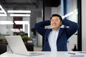 Satisfied with the result, a young Asian male businessman sits relaxed in a chair at a desk in the...