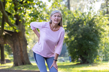 An elderly gray-haired woman felt severe back pain while walking and jogging in the park, standing...