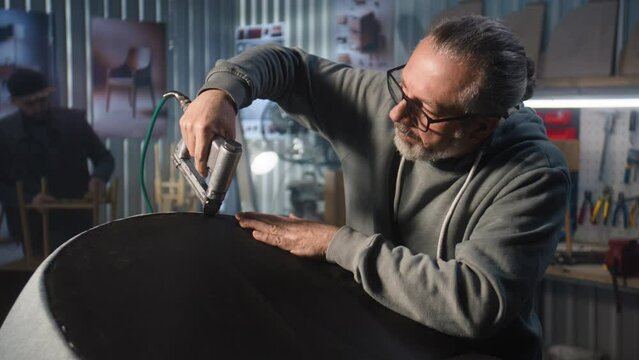 Mature carpenter makes upholstery of chair using staple gun in stylish workshop for making and restoration furniture. Male colleague assembles shelf in the background. Handcraft and small business.