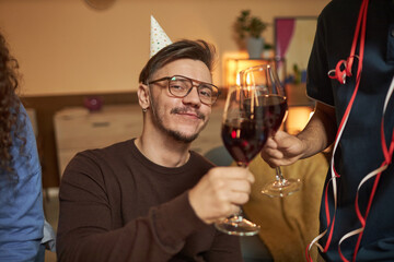 Close up portrait of smiling man with disability wearing party hat and clinking wine glasses with friends while smiling at camera
