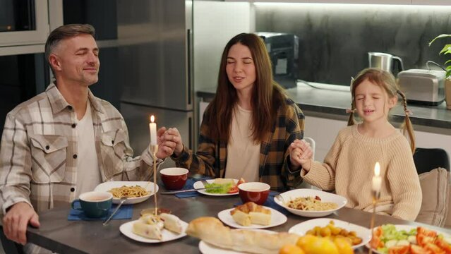 Happy family brunette woman in a plaid shirt together with her middle aged husband with gray hair and little daughter praying and giving thanks for the food before starting an evening dinner in a