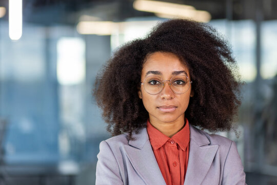 Close-up portrait of a serious and successful young ,African American woman in a suit and glasses standing in the office and looking smugly at the camera.