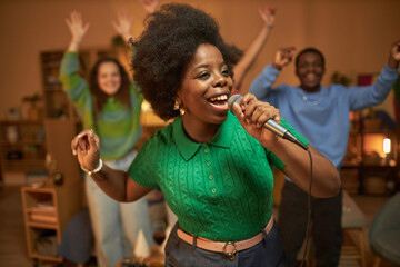 Waist up portrait of carefree African American woman singing to microphone and dancing enjoying karaoke at home party