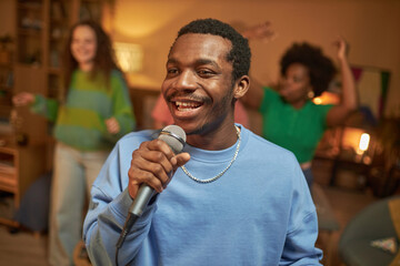 Waist up portrait of Black adult man singing to microphone and smiling happily enjoying karaoke at...