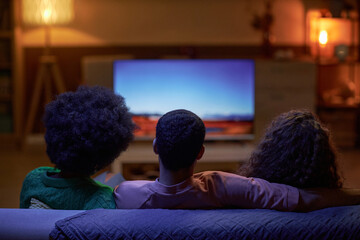 Back view of three people sitting on couch together and watching TV in dark room