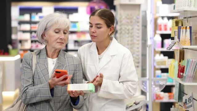 Concerned old woman showing to young female pharmacist QR-code of product on her smartphone in chemist's shop. High quality 4k footage