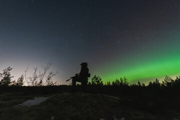 Fototapeta premium A military soldier with a night vision device and a rifle with a silencer is sitting in the forest against the background of the starry sky and northern lights.