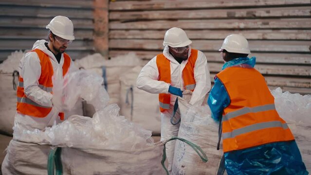 A team of three workers in a white uniform and an orange vest stack and press polyethylene and cellophane while working at a large waste recycling plant