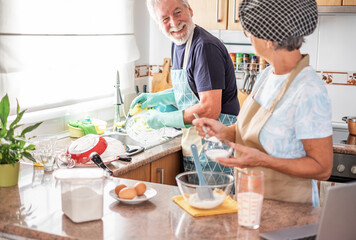 Happy senior couple working together in home kitchen, husband washes dishes, wife prepares homemade...