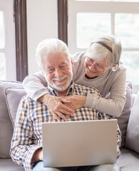 Happy white-haired 70-year-old couple at home using laptop together to shop online, husband sits on sofa, wife hugs him. Older people use new technologies