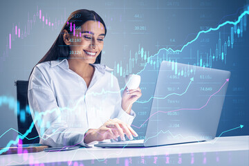 Attractive happy businesswoman using laptop at desk with creative glowing candlestick forex chart...