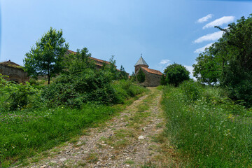 Dirt road to the church. The stone wall and dome are visible. Trees and bushes around. Bright blue sky with clouds
