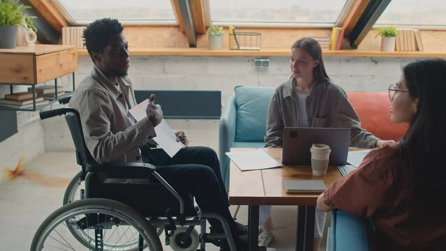 Medium shot of African American man with disability sitting in wheelchair in loft coworking office at project team meeting, proposing new business idea and showing graphs, female colleagues listening