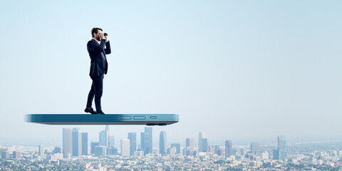 A focused businessman uses binoculars atop a smartphone, overlooking the city, representing vision...