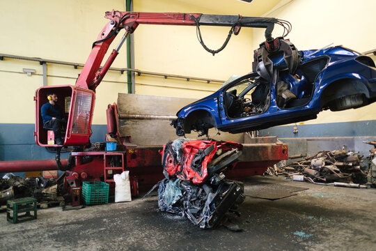 Mechanic operating crane machine and carrying scrapped car at workshop