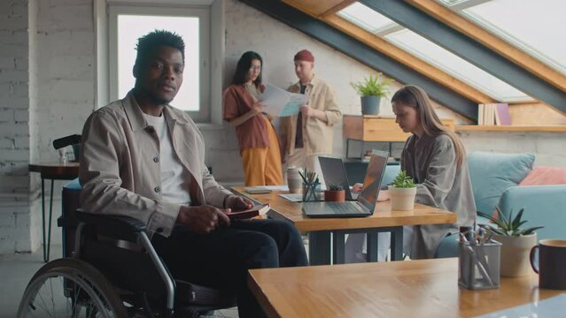 Medium full portrait shot of African American man with disability sitting in wheelchair in loft shared office facility, closing notebook and looking on camera, other team members working in background