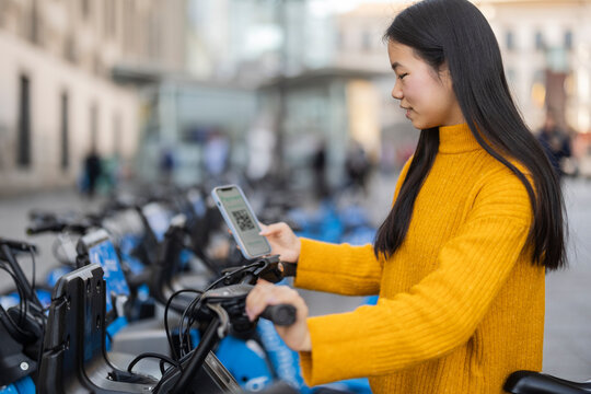 Young woman scanning QR code through smart phone and renting electric bicycle