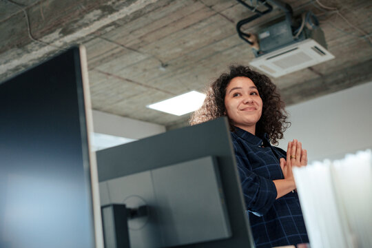 Smiling businesswoman standing with hands clasped near computer in office