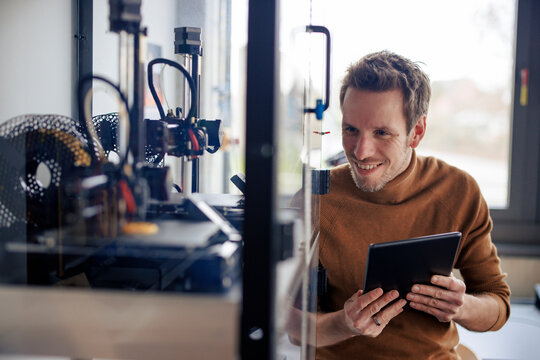 Smiling engineer holding tablet PC and looking at 3d printer in workshop
