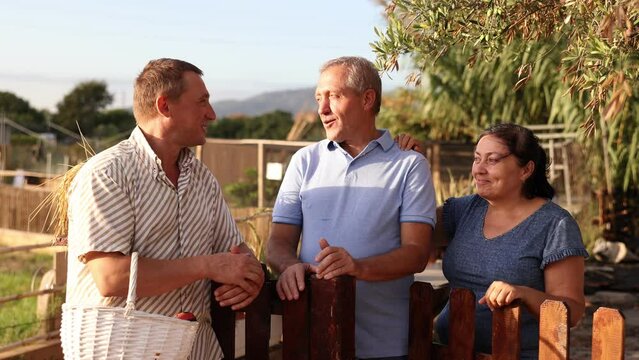Couple of smiling elderly amateur gardeners discussing interesting topics with their male neighbor while standing near wooden fence in garden on summer day