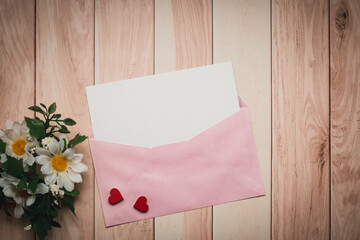 A pink envelope and a white card with a red heart and daisy are placed on a wooden table