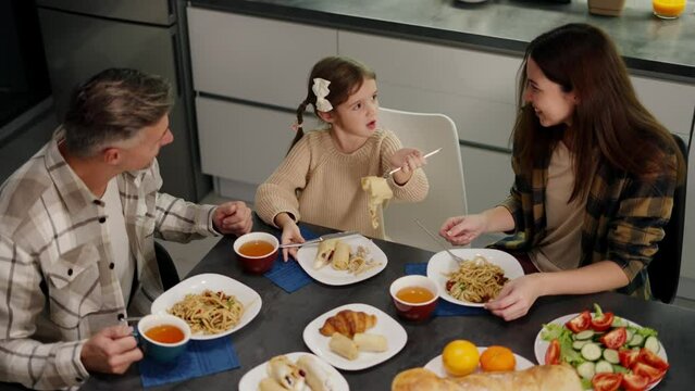 Top view of a happy family having dinner together at the table in the kitchen in the evening. Happy little girl with her brunette mom in a plaid shirt and middle-aged dad and gray hair having dinner