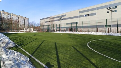 The stadium is located among various buildings and trees. The soccer field with goals and artificial grass is cleared of snow. Pillars with lighting lanterns are installed around it. Sunny weather