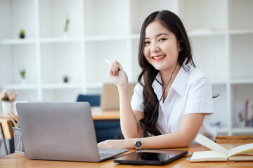 Happy young asian woman wearing earphones and using laptop computer at desk in office, Female asian student with laptop computer, College female student learning online.
