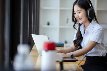 Happy young asian woman wearing earphones and using laptop computer at desk in office, Female asian...