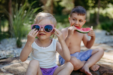 Little chidren sitting near lake and eating watermelon on hot sunny day during summer vacation.