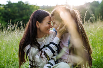 Beautiful mother with daughter, touching with foreheads, sitting in the grass at meadow. Concept of...