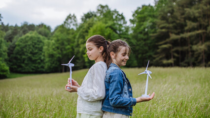Girls holding model wind turbine, standing on meadow, in nature. Concept of renewable, green energy and sustainable lifestyle for future generations.