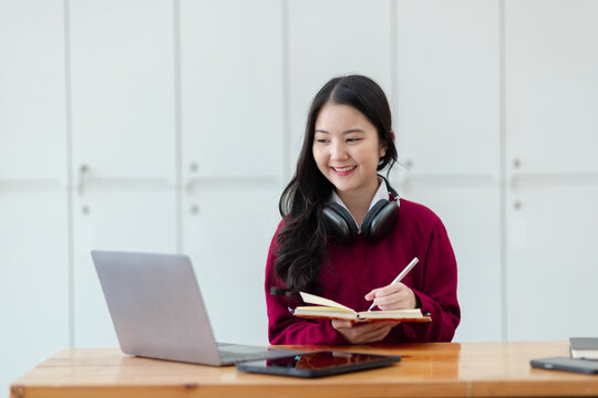 Happy young asian woman wearing earphones and using laptop computer at desk in office, Female student with stack of books and laptop.
