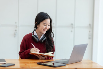 Happy young asian woman wearing earphones and using laptop computer at desk in office, Female student with stack of books and laptop.