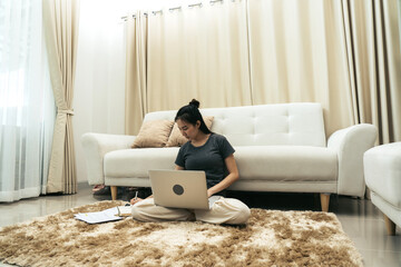 Asian woman sits on a rug in the living room and works on a computer while writing down notes in a notebook.