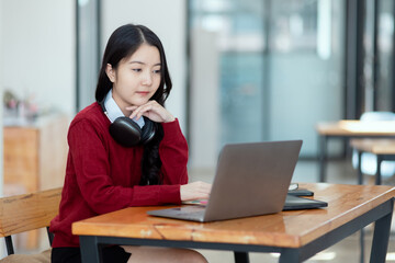 Happy young asian woman wearing earphones and using laptop computer at desk in office, Female student with stack of books and laptop.
