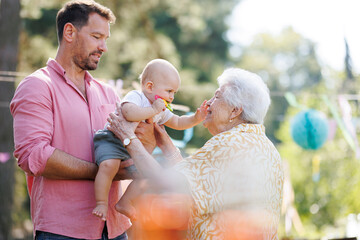 Great-grandmother holding little baby in her arms. Family summer garden party. - 750471915