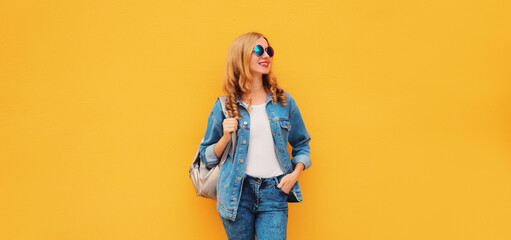 Portrait of stylish modern smiling young woman with backpack in denim jacket on yellow background