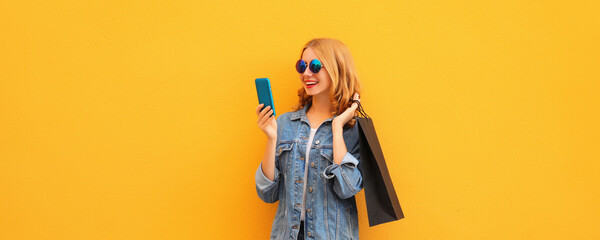 Beautiful happy young woman looking at mobile phone with black shopping bag on yellow background