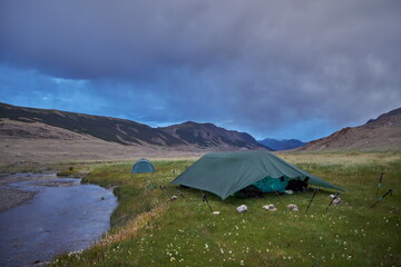 Camping tents nestled in a wild meadow by a stream with looming mountains under a stormy sky