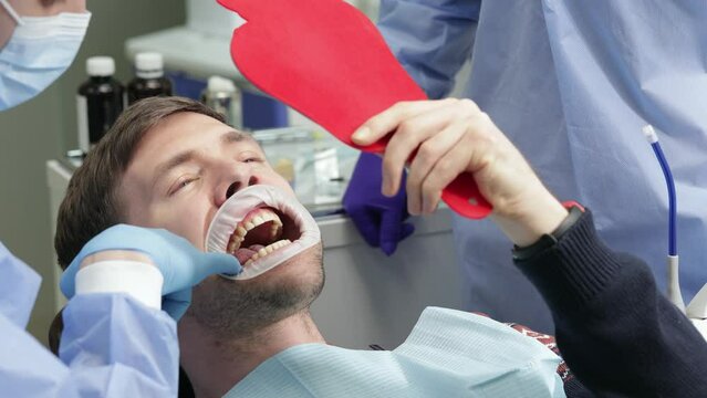 Man with cheek retractor looking at mirror enjoy healthy clean teeth beautiful smile after treatment at stomatology clinic. Handsome male patient and dentist doctor admiring result tooth whitening