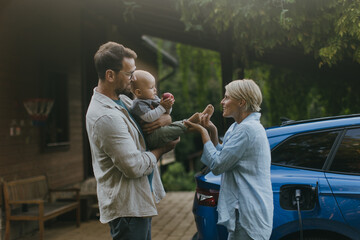 Young family with baby standing by their electric car. Electric vehicle with charger in charging...