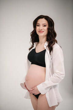 Pregnant woman holds belly stands near a white wall. Young woman on a light background. Pregnancy motherhood concept. Female waiting for baby. Pregnant girl touching belly, caring health indoors.